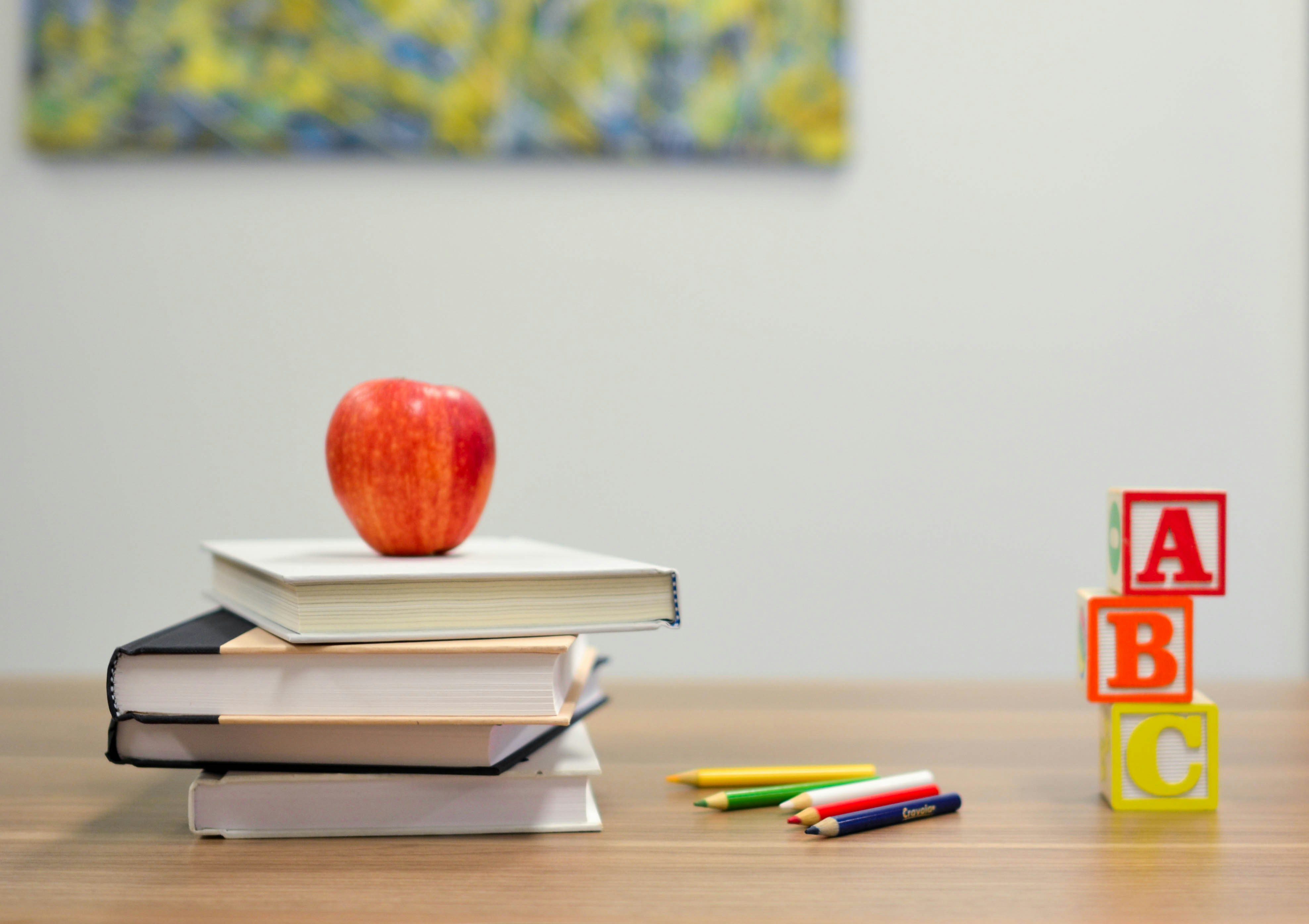 A red apple rests on stacked books next to colored pencils and building blocks labeled A, B, and C on a wooden table.